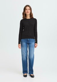 Cardigan noir côtelé avec fermeture à boutons, manches longues, et design ajusté, associé à un jean évasé bleu clair et des chaussures plates noires.