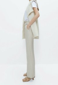 Beżowe spodnie w wzór houndstooth o tapered fit, zestawione z teksturowanym kremowym kamizelką i podstawowym białym t-shirtem, na neutralnym tle.