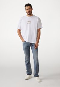 Hvid bomulds T-shirt med et trykt logo i beige. Kombination med lyseblå denim jeans og hvide sneakers. Casual pasform, korte ærmer.
