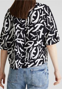 Blouse noire à manches courtes avec un motif abstrait blanc. Coupe ample avec un ourlet arrondi. Fabriquée en tissu lisse, assortie à un jean bleu clair.