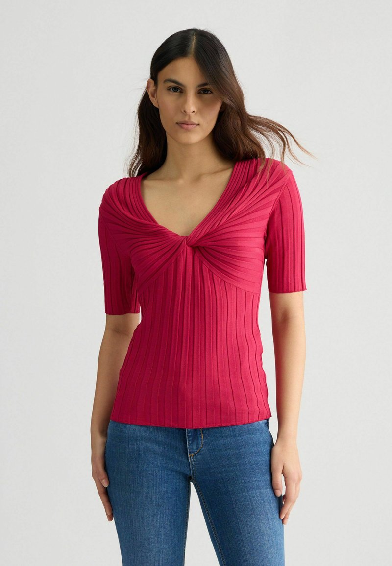 Tricou scurt, ajustat, de culoare roz aprins, cu decolteu adânc în V, textură plisată și model cu dungi, asortat cu blugi albaștri.