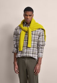 Grün-beige kariertes Hemd mit langen Ärmeln, kombiniert mit einem leuchtend gelben Strickpullover, der um den Hals drapiert ist, und olivgrünen Hosen.