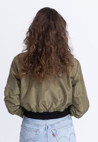 Giacca bomber corta verde oliva con maniche a sbuffo, caratterizzata da una vita a coste nere e una texture liscia. Indossata con jeans chiari.