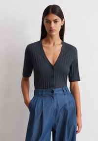 Czarny, prążkowany sweter z głębokim dekoltem w serek i krótkimi rękawami, z rządem czarnych guzików na przodzie; zestawiony z niebieskimi spodniami w kant.