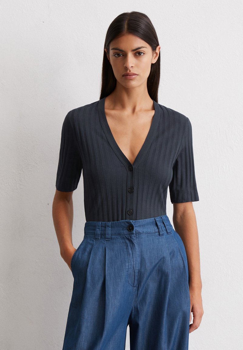 Czarny, prążkowany sweter z głębokim dekoltem w serek i krótkimi rękawami, z rządem czarnych guzików na przodzie; zestawiony z niebieskimi spodniami w kant.