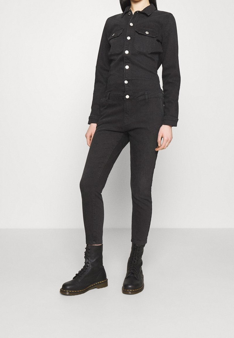 Salopette en denim noir avec un devant à boutons, deux poches poitrine et une silhouette ajustée. Assortie à des bottes de combat noires à lacets.