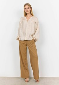 Lys beige, knap-op blouse med plisseret tekstur og lange ærmer. Parret med højtaljede, brede brune bukser, der fremhæver en afslappet pasform.