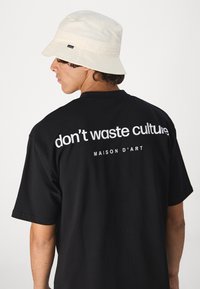 Camiseta negra con texto blanco en la parte posterior que dice "no desperdicies la cultura" y "MAISON D'ART." Complementada por un sombrero tipo bucket beige.