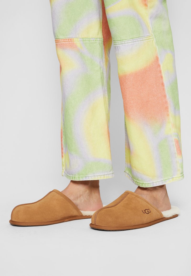 Braune Wildlederhausschuhe mit einem weichen Innenfutter und abgerundetem Zeh, versehen mit einem Logo an der Seite, kombiniert mit mehrfarbigen Batik-Hosen.