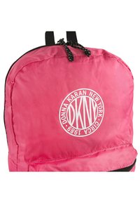 Mochila de nylon rosa con un logo redondo blanco que dice "Donna Karan New York" y detalles en negro en los cierres. Correas ajustables negras en la parte superior.
