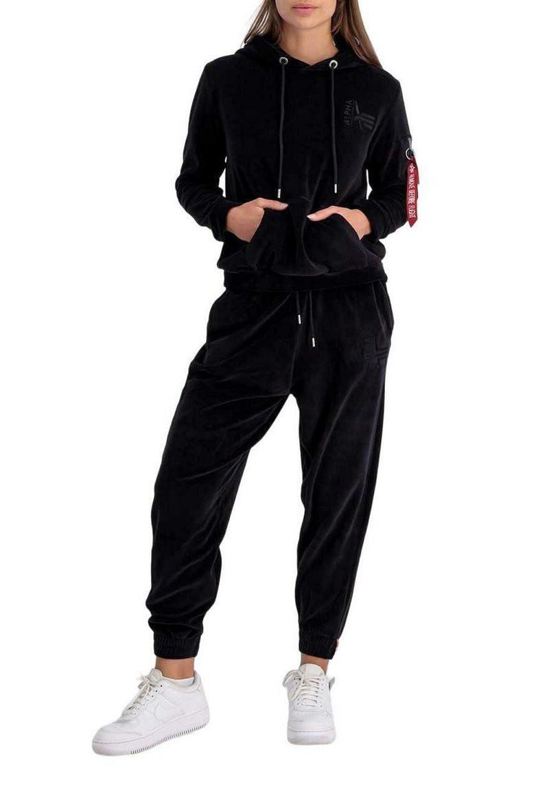 Schwarzes Velours-Hoodie und Jogginganzug-Set mit einer Fronttasche, Kordelzügen und Seitentaschen. Weiße Turnschuhe vervollständigen das Outfit.