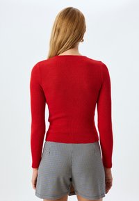 Rød ribbestrikket sweater med lange ærmer, med en rund halsudskæring og tætsiddende pasform, parret med sorte og hvide ternede shorts.