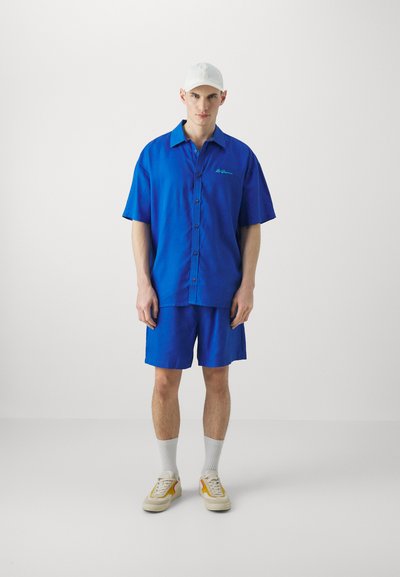 Koszula na krótki rękaw zapinana na guziki i dopasowane szorty w jasnoniebieskim kolorze, z zielonym logo. W połączeniu z białą czapką i beżowymi butami.