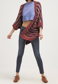 Cárdigan de punto con un patrón de rayas rojas y azules, con apertura frontal y mangas anchas, combinado con un suéter corto azul y jeans oscuros.
