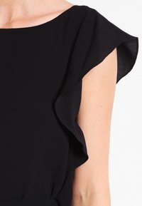 Manche de robe noire à volants en tissu léger. Texture lisse avec une silhouette douce et fluide et un décolleté large. Pas d'ornements.