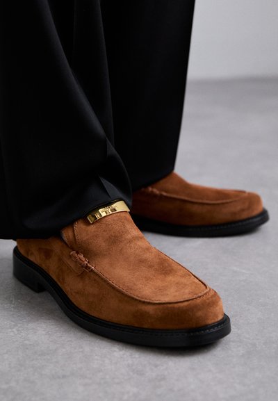 Mocassins em camurça castanha com uma faixa dourada, apresentando biqueira redonda e sola preta plana. Usados com calças pretas.