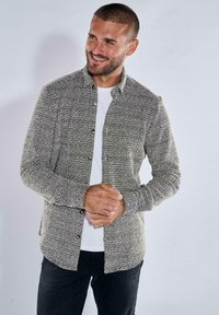 Grau- und schwarz gemustertes Hemd mit klassischem Kragen, langen Ärmeln, strukturiertem Stoff und Knopfverschluss, über einem weißen Hemd getragen.