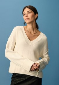 Creme V-hals sweater med ribbet tekstur og oversized ærmer, parret med en sort lædernederdel. Baggrunden er ensfarvet blå.