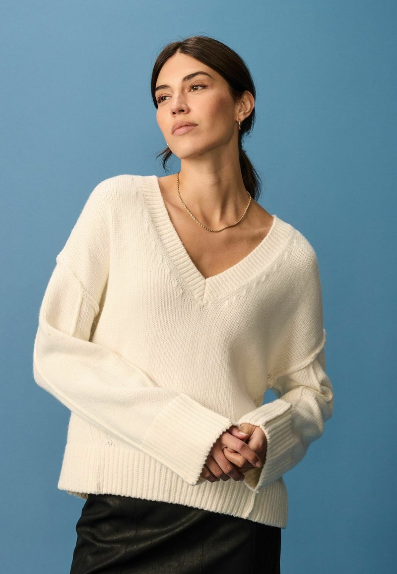 Creme V-hals sweater med ribbet tekstur og oversized ærmer, parret med en sort lædernederdel. Baggrunden er ensfarvet blå.