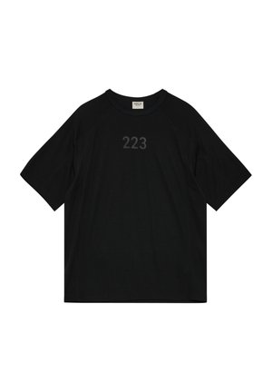 Mann, der frontal steht und ein schwarzes übergroßes T-Shirt mit der Aufschrift „223“ auf der Brust, graue lockere Hose und graue Sneaker auf weißem Hintergrund trägt.