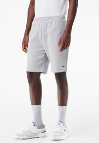 Homme portant un short gris clair à taille élastique avec un petit logo crocodile vert, des chaussettes blanches, et des baskets blanches avec des accents bleus.