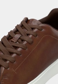Cauri brūni ādas sporta apavi ar teksturētu virsmu, aprīkoti ar plakanu brūnu šņorēm un baltu gumijas zoli. Tuvplāns uzsver izšūšanas detaļas.