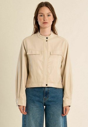 Giovane donna con capelli castani lisci che indossa una giacca beige chiaro con bottoni e jeans di denim blu, in piedi davanti a uno sfondo semplice.