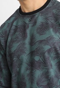 Felpa verde con motivo di foglie di palma nere, collo a costine e tessuto morbido, dal taglio casual e rilassato.