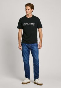 Melns kokvilnas T-krekls ar baltu "PEPE JEANS" logo, kombinēts ar ziliem džinsiem. Modelis valkā baltas sporta kurpes, stāvot pret vieglu fona.