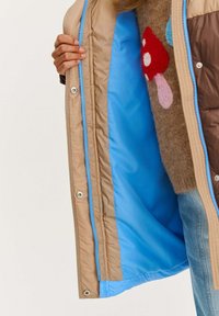 Osoba drží otvorenú béžovú bundu s modrým podšívkou, na sebe má hnedý sveter s farebnými vzormi húb a svetlomodré džínsy.