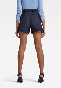 Námořnické modré šortky s ohrnutými manžetami, elastickým pasem a zadními kapsami, s nápadným prošíváním pro větší detail. Hladká struktura tkaniny.