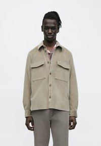 Lichtbeige corduroy overhemdjas met twee borstzakken, knoopsluiting en een ontspannen pasvorm. Soepele textuur met een klassieke kraag.