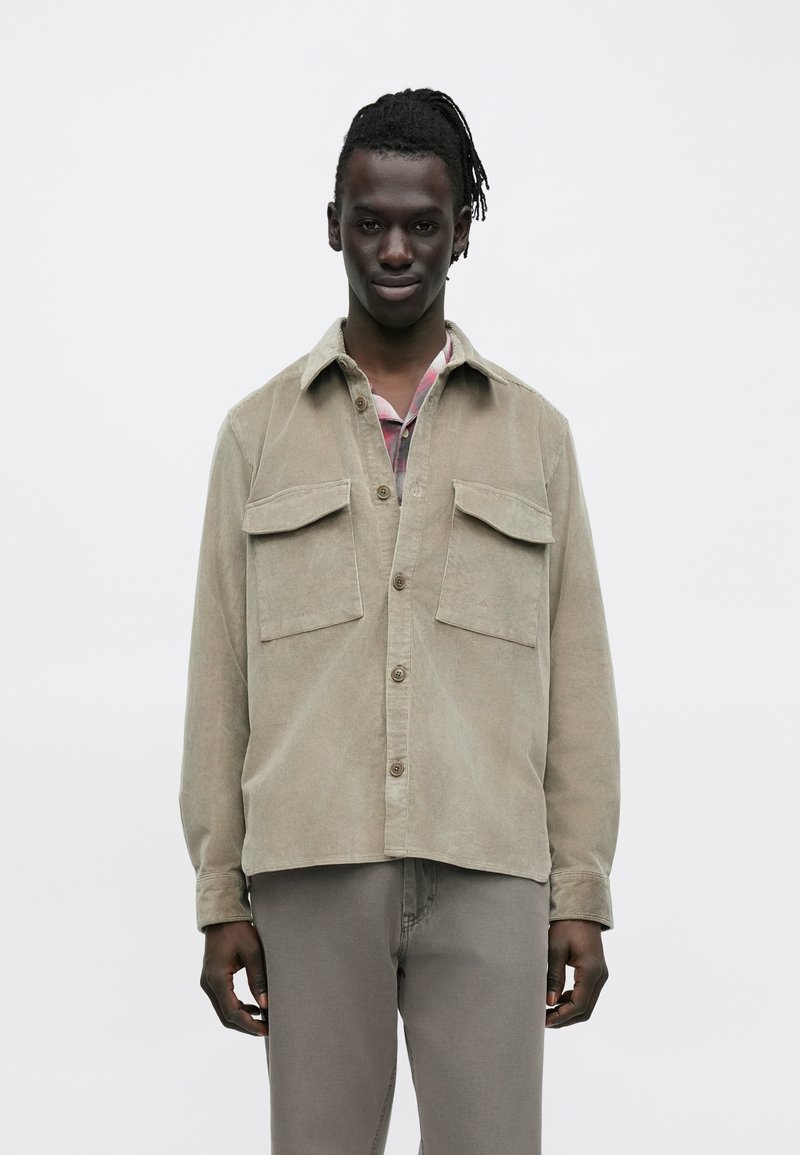 Lichtbeige corduroy overhemdjas met twee borstzakken, knoopsluiting en een ontspannen pasvorm. Soepele textuur met een klassieke kraag.