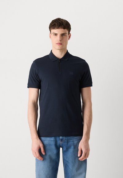 Polo shirt bleu marine avec col zippé, manches courtes et un petit écusson logo, fabriqué dans un tissu lisse avec une coupe ajustée.