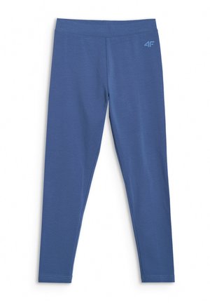 Młoda dziewczyna ubrana w różową czapkę typu beanie, kremową bluzę zapinaną na suwak, niebieskie legginsy, białe skarpetki i białe trampki, stoi na tle jednolitego jasnego tła.