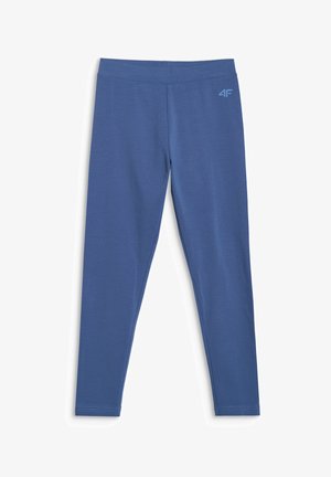 Jeune fille portant un bonnet rose, un sweat zippé crème, des leggings bleus, des chaussettes blanches et des baskets blanches, debout devant un fond clair uni.