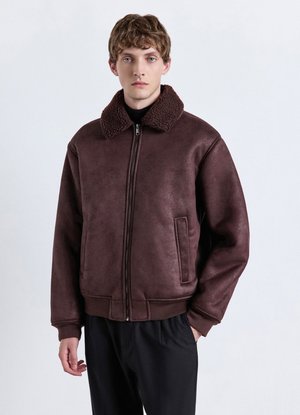 Junger Mann trägt eine dunkelbraune Reißverschlussjacke mit dickem Schaffellkragen und schwarze Hose, steht vor einem einfarbigen hellen Hintergrund.