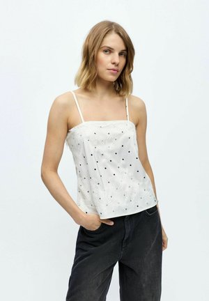 Jeune femme aux cheveux blonds courts portant un haut sans manches blanc brodé avec des pois noirs et un pantalon noir, debout avec une main dans la poche.