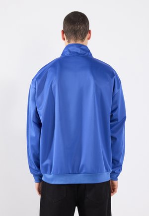 Man die wegkijkt en een felblauwe lange mouwen jas met een hoge kraag en zwarte broek draagt tegen een effen achtergrond.