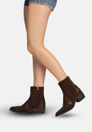 Benen met donkergroene, textuurrijke enkellaarsjes met zijrits en blokhakken, gecombineerd met denim shorts op een witte achtergrond.