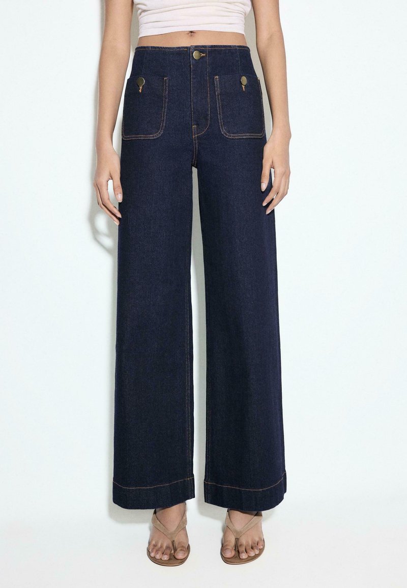 Femme portant un jean taille haute, jambes larges, bleu foncé, avec poches plaquées boutonnées à l'avant et des sandales beiges, top blanc court partiellement visible.