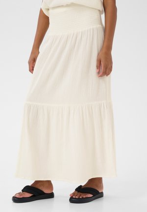 Femme debout portant une longue jupe crème avec une taille smockée et des sandales noires à enfiler sur un fond clair uni.