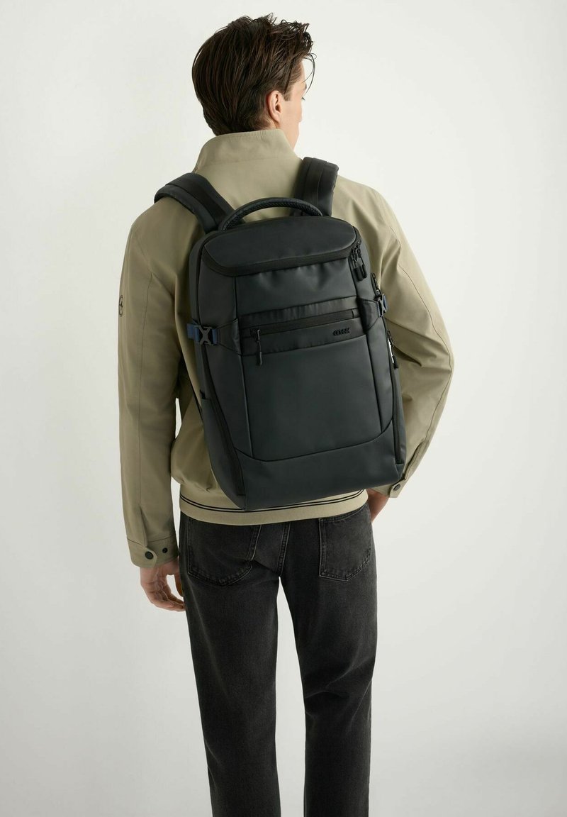 Mężczyzna w beżowej kurtce i czarnych dżinsach, niosący czarny plecak z wieloma przegródkami, stojący na tle jasnego, jednolitego tła.