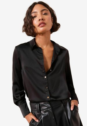 Sort satin langærmet skjorte med krave og knaplukning foran. Stoffet har en glat tekstur og er parret med høj talje læderbukser.