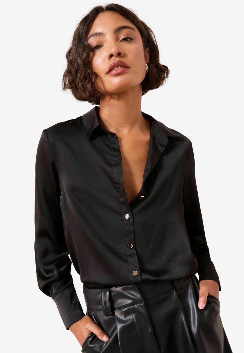 Sort satin langærmet skjorte med krave og knaplukning foran. Stoffet har en glat tekstur og er parret med høj talje læderbukser.