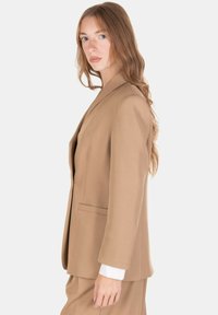 Blazer color cammello con una vestibilità su misura, rever a taglio, tasche frontali e texture liscia. Indossato sopra una blusa bianca.