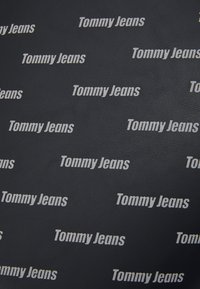 Černý texturovaný povrch s několika bílými logy "Tommy Jeans" rovnoměrně rozmístěnými, které zobrazují opakující se vzor značky.