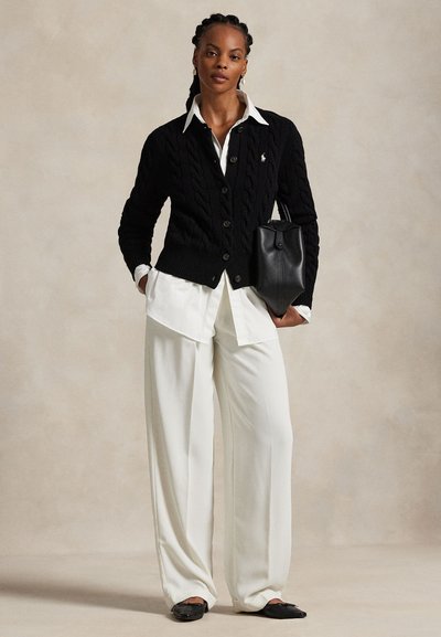 Fonott hajú nő fekete, kötött mintás kardigánt visel fehér ing fölött, fehér, bő szárú nadrágban, kezében fekete bőr kézitáskát tart.