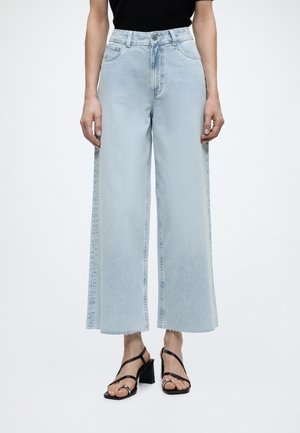 Άτομο φοράει ανοιχτό μπλε τζιν παντελόνι φαρδιές cropped γραμμής και μαύρα πέδιλα με τακούνι και λουράκια, στέκεται μπροστά σε απλό λευκό φόντο.