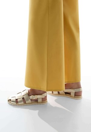 Sandales strappy beige clair avec détail à boucle, semelle texturée et tige tissée, associées à un pantalon jaune à jambes larges.